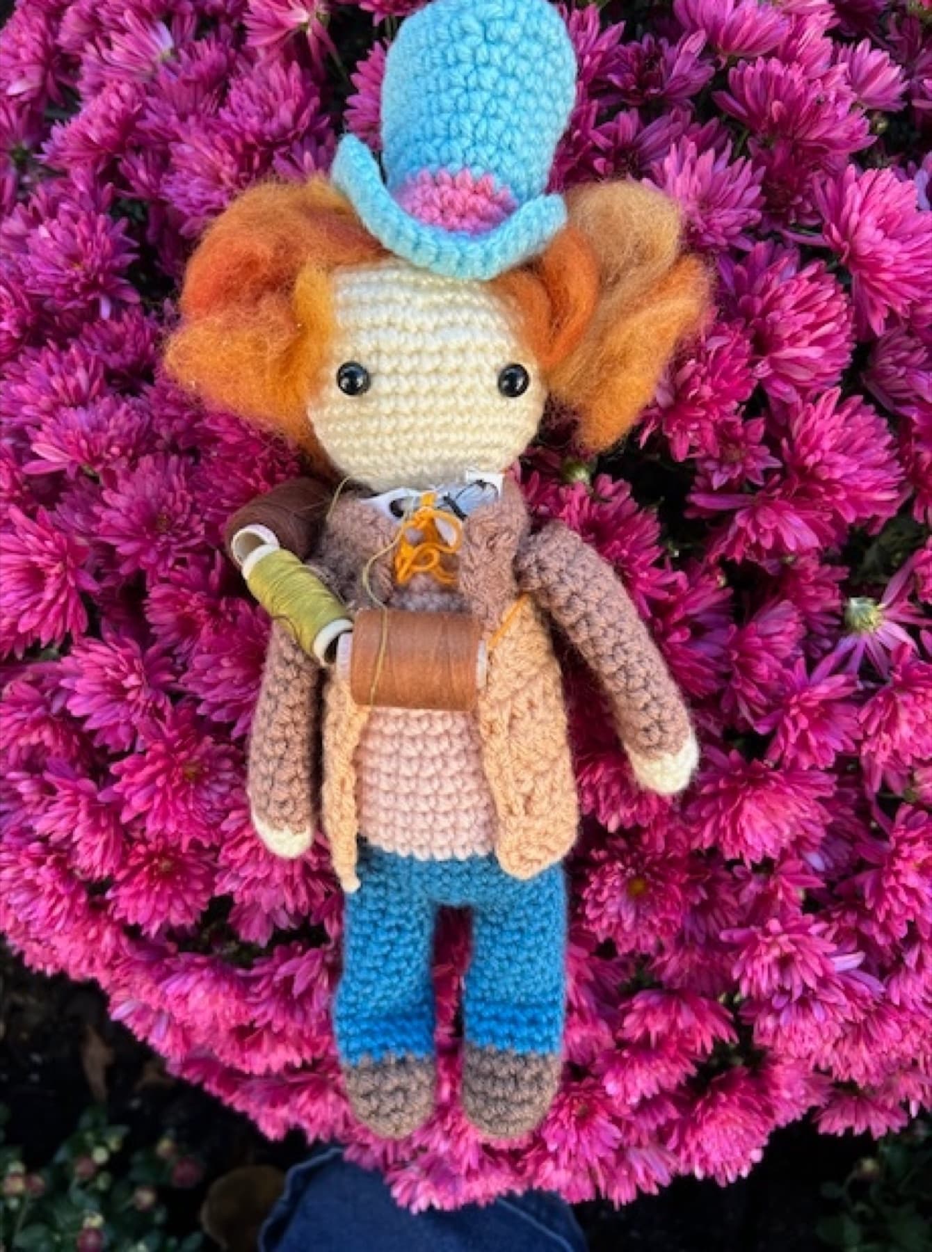 A crocheted doll with orange wool hair and a blue hat, carrying tiny spools of thread, resting on magenta chrysanthemums