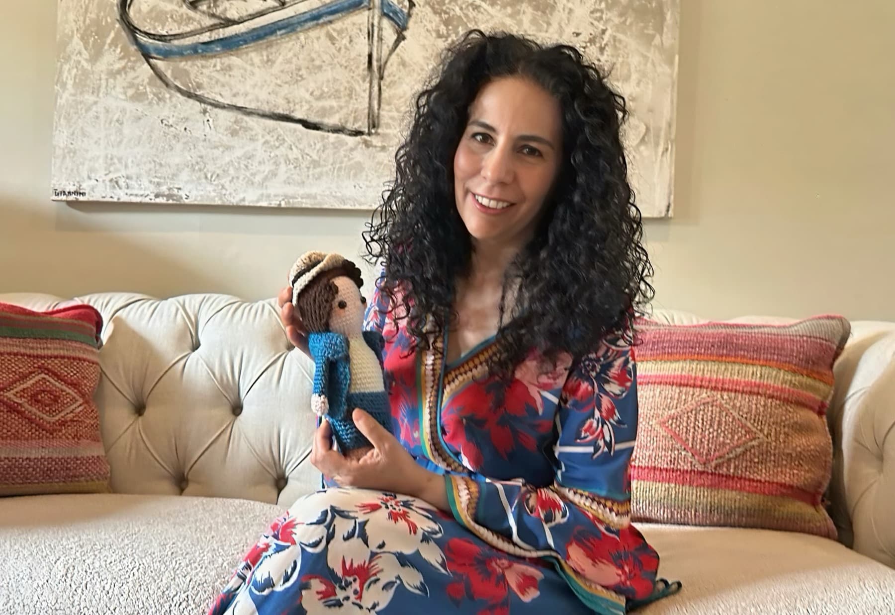 Maria smiling on a cream sofa, holding a small crocheted doll in a blue outfit and hat