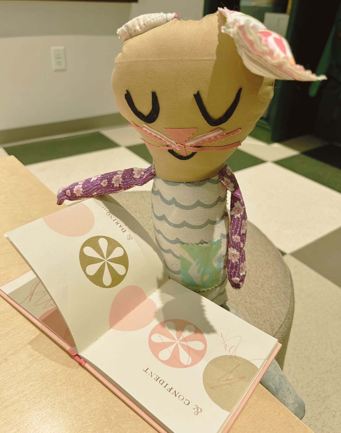 A sewn cat toy with fabric ears, cherry-blossom print arms, a wave-pattern body, and ric-rac whiskers, reading a picture book