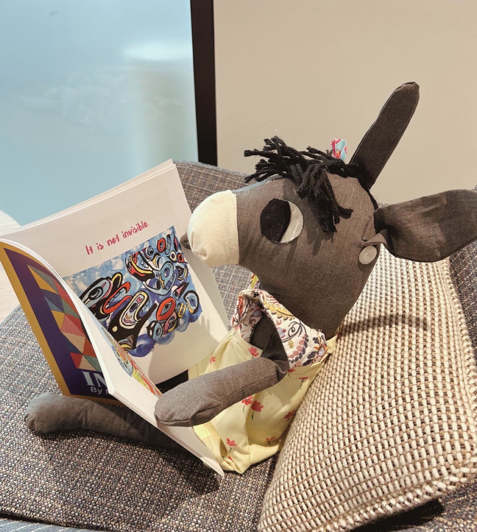 A sewn gray donkey toy with a black yarn mane, a button ear, and a yellow floral dress, reading an art book