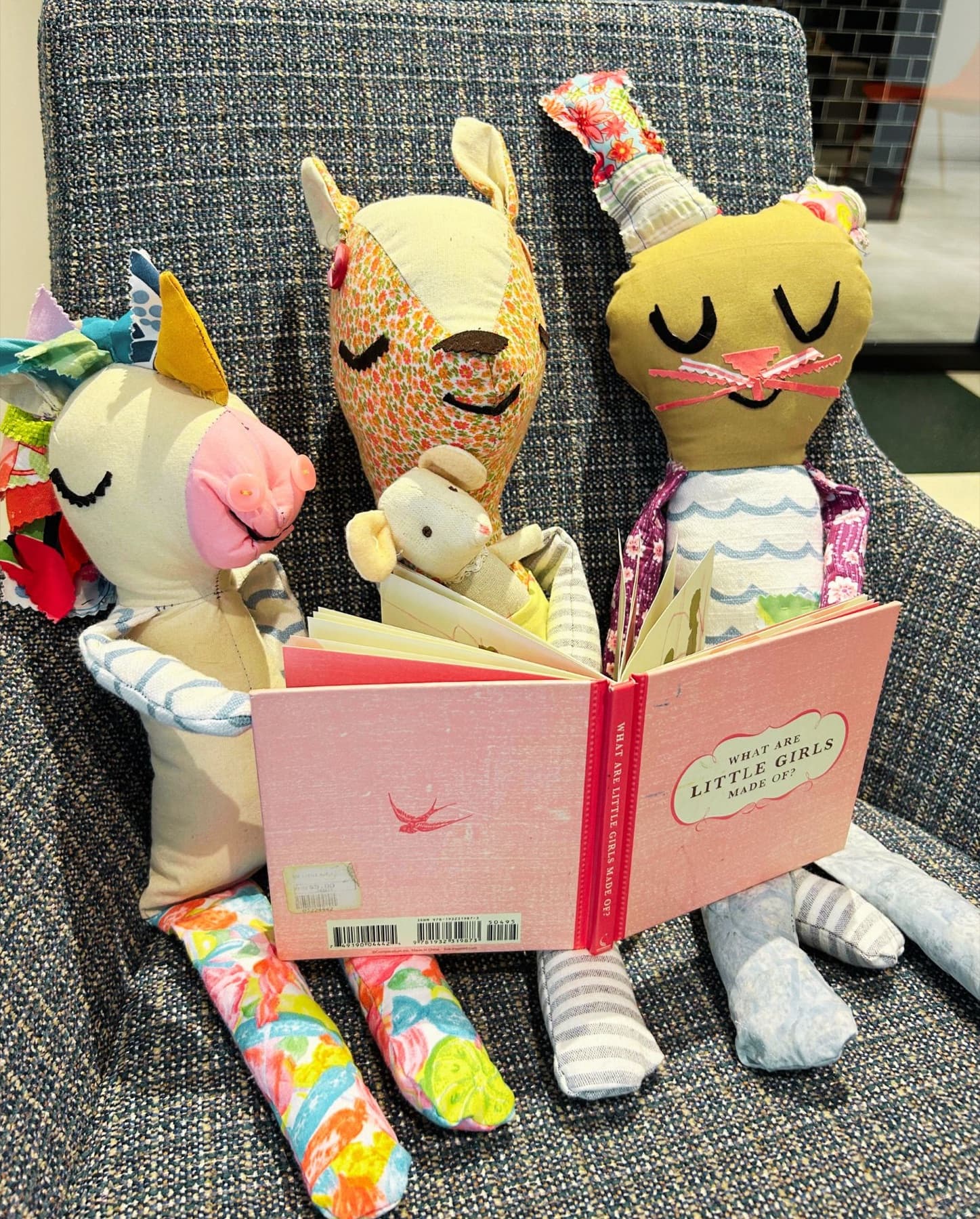 Three handmade fabric toys — a patchwork unicorn, a floral deer holding a tiny mouse, and a cat — sitting on a tweed chair sharing a pink picture book